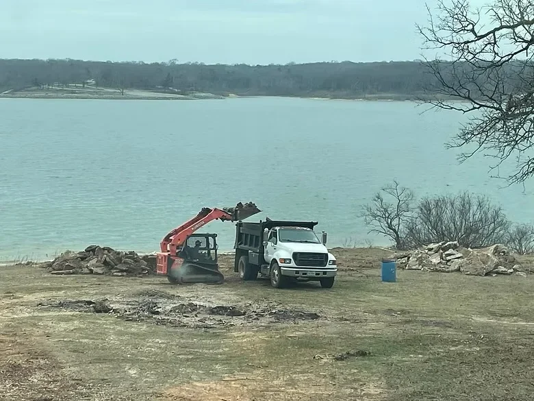 Tracked excavator loading dump truck with excavated soil and rock at waterfront site