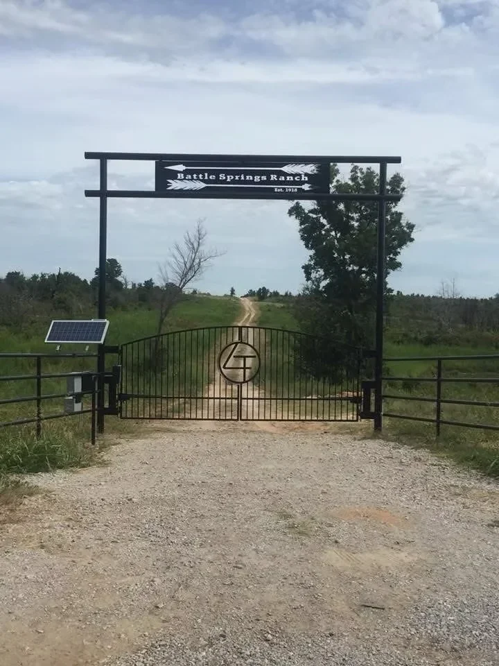 Black steel ranch entry gate with logo cutout and overhead sign structure on gravel driveway