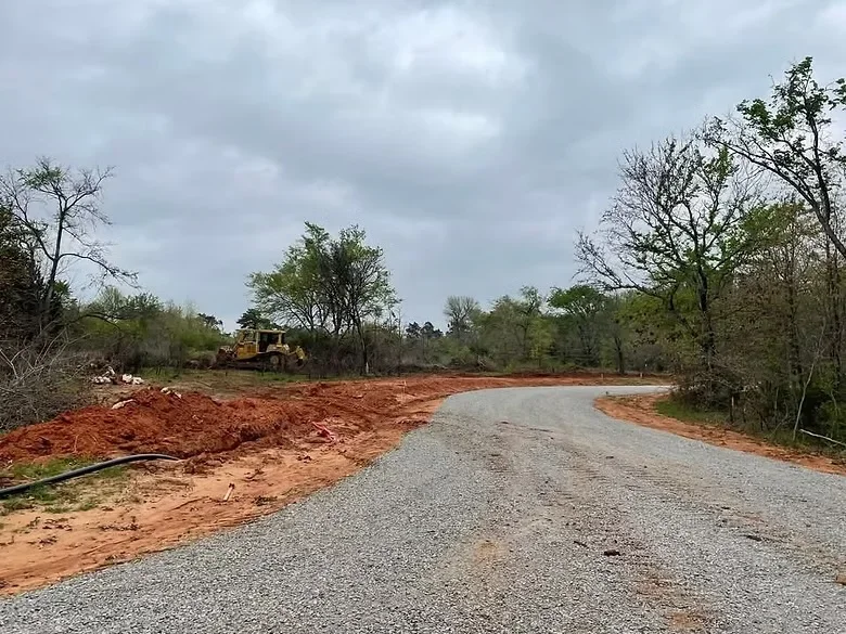 Graded driveway base with compacted gravel and red clay soil, heavy equipment visible on cleared property