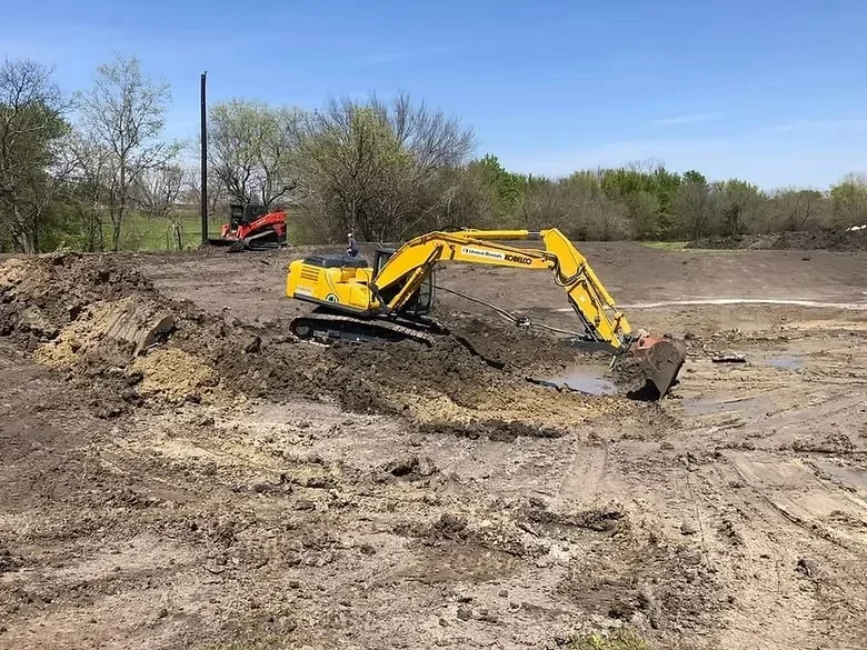Yellow excavator digging and grading dirt lot with red compactor in background