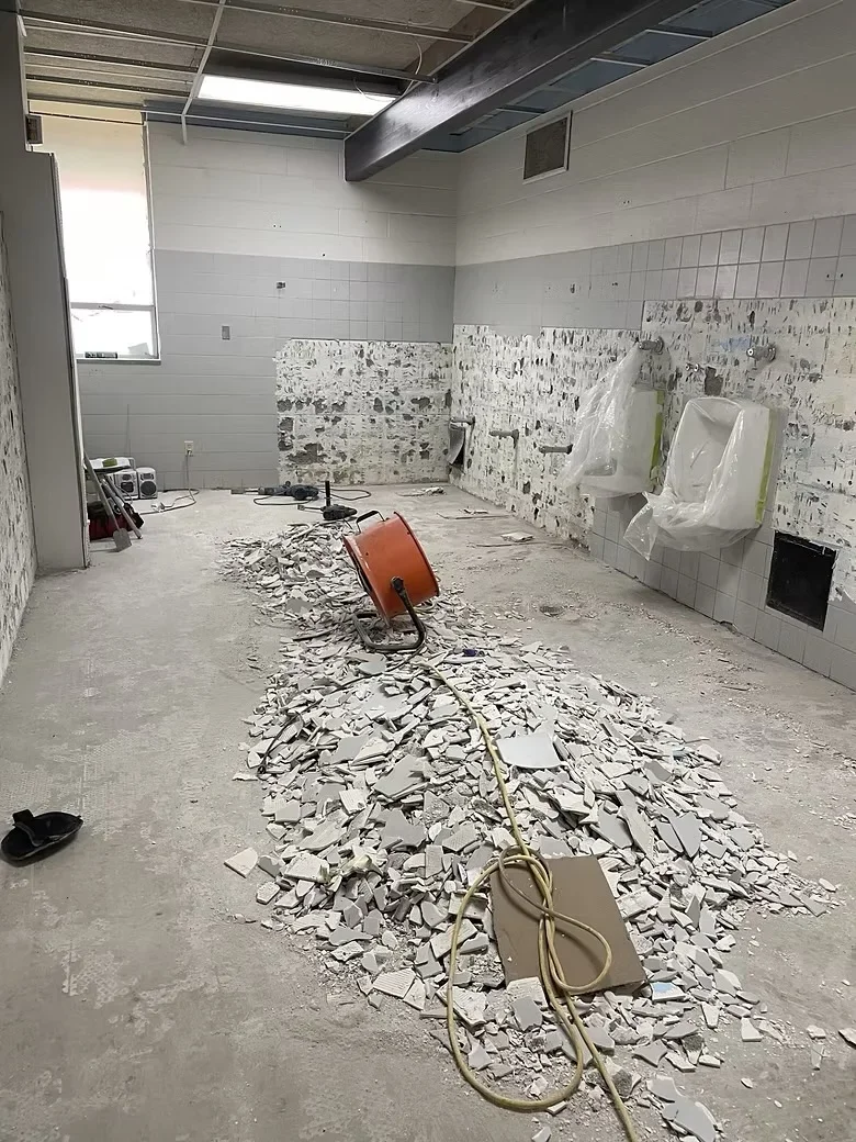 Commercial restroom interior with tile removal debris, exposed concrete floor, and protective coverings on remaining fixtures