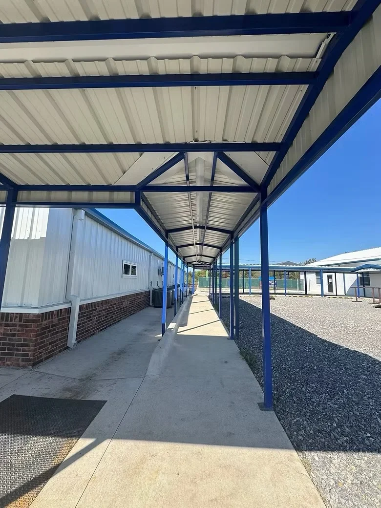 Steel canopy with blue beams and white metal roofing over concrete walkway and pad