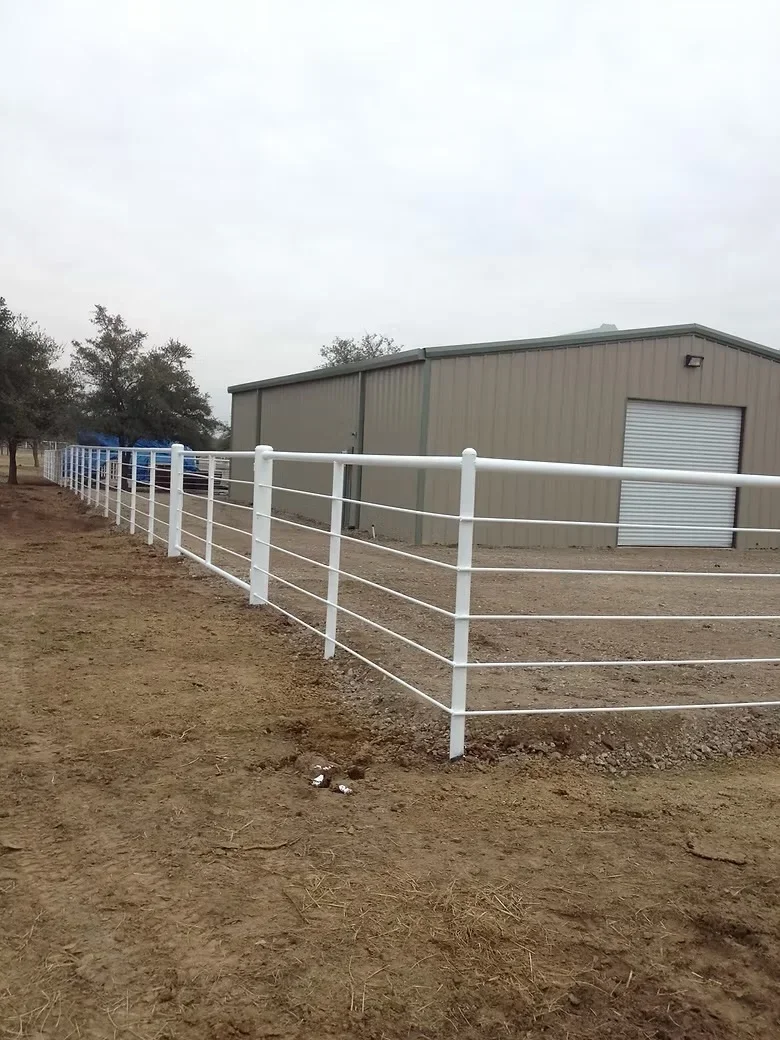 White metal pipe rail fence with four horizontal rails installed on dirt ground near metal agricultural building