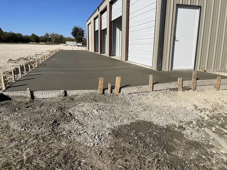 Finished concrete pad with troweled surface and wooden grade stakes beside metal building