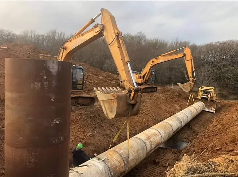Excavators positioning concrete utility pipe in trench with earthwork and safety personnel